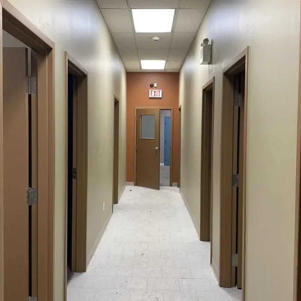 A long, narrow hallway with several doors on either side and a door at the end leading to an exit.
