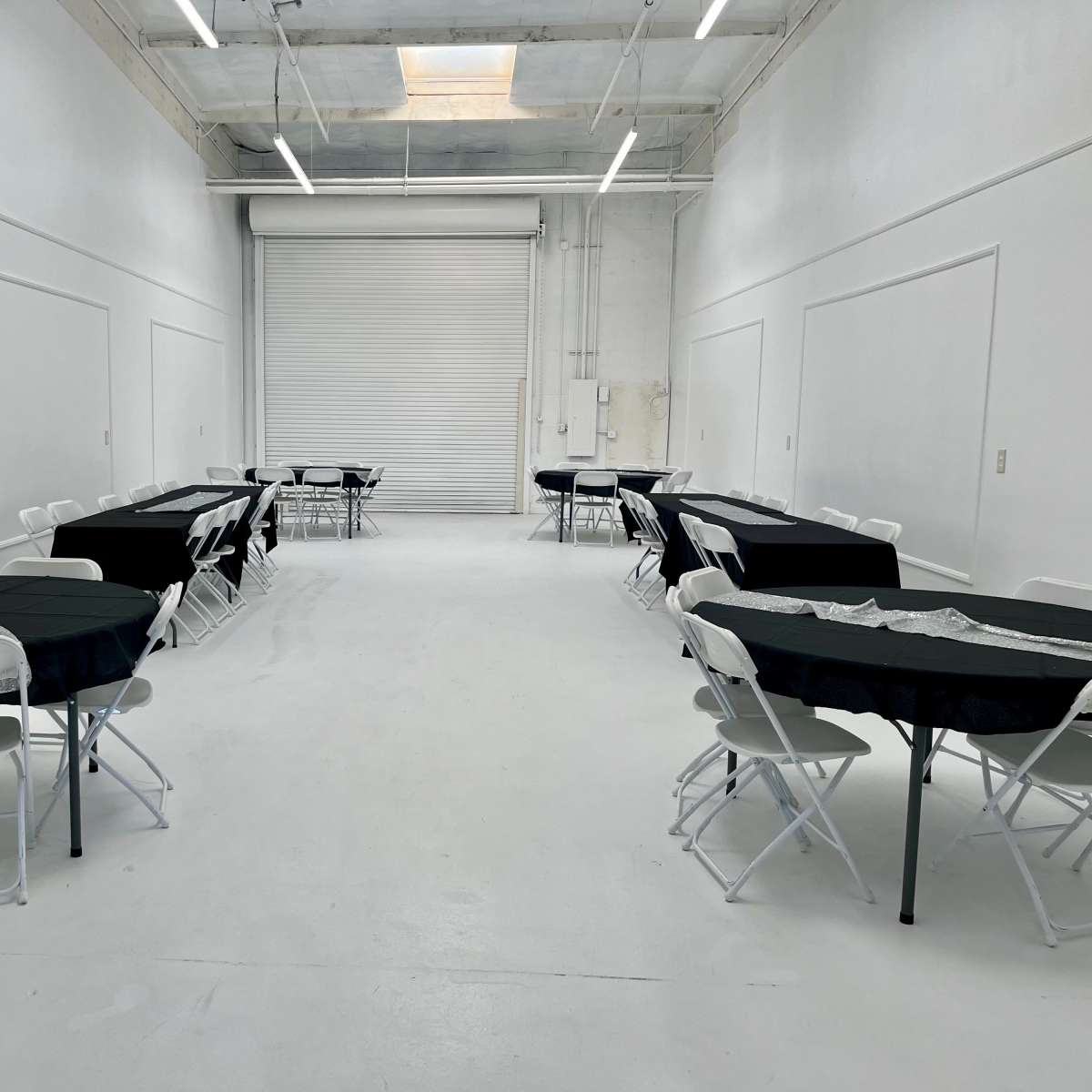 A spacious, white room with multiple round tables covered in black tablecloths and surrounded by folding chairs, arranged in two rows.