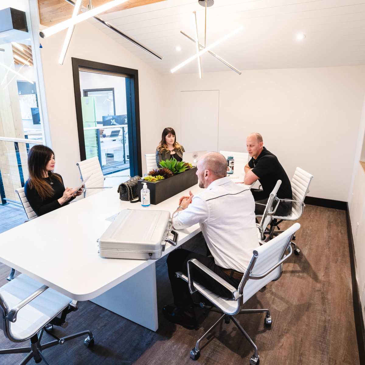 A group of four people is seated around a modern conference table in a well-lit meeting room with green wall art.