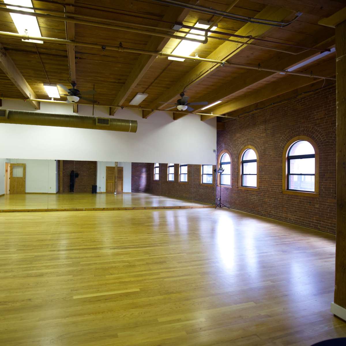 An empty dance studio with wooden floors, exposed beams, and large arched windows.