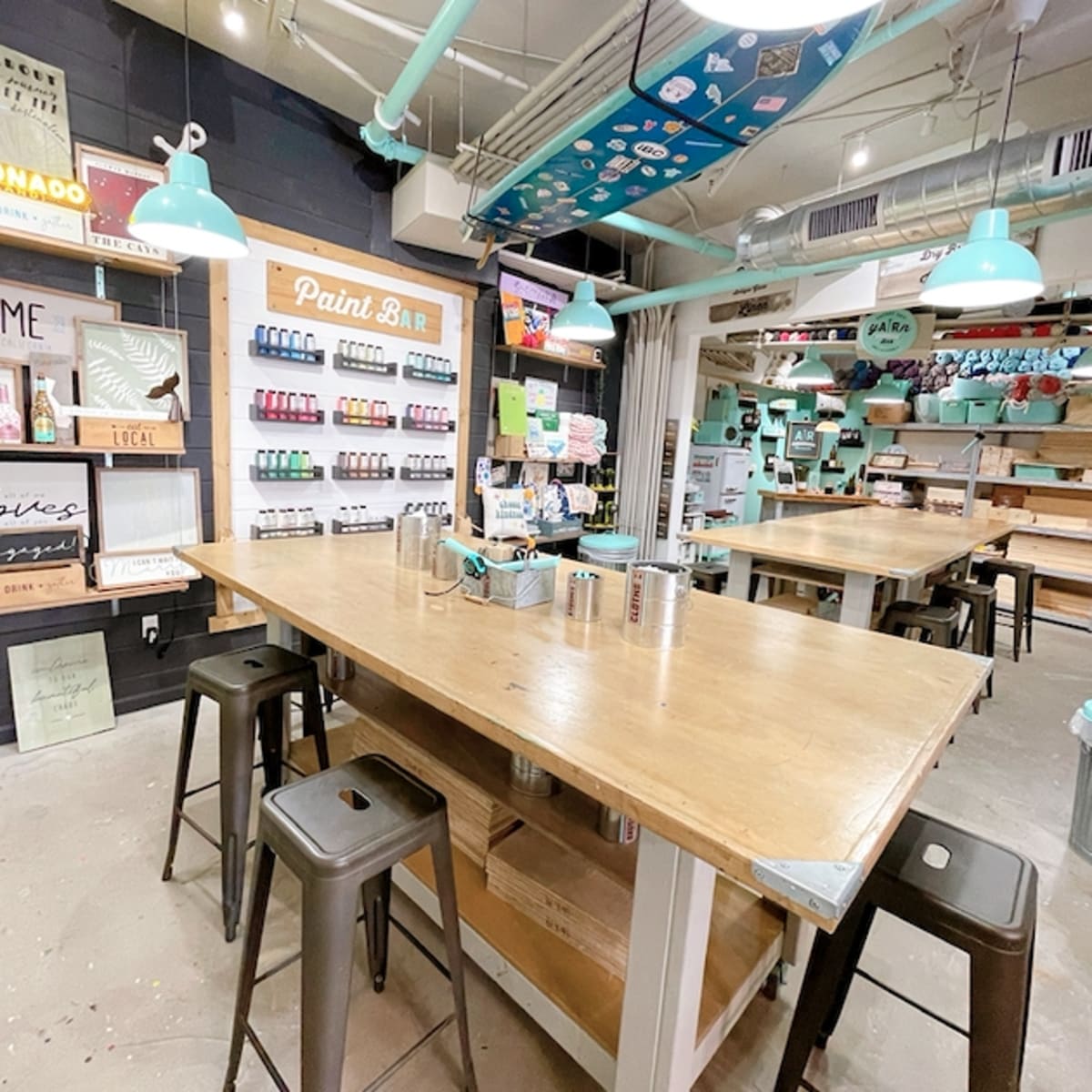 A brightly lit art studio with several wooden tables, teal pendant lights, and shelves displaying an assortment of paint supplies.