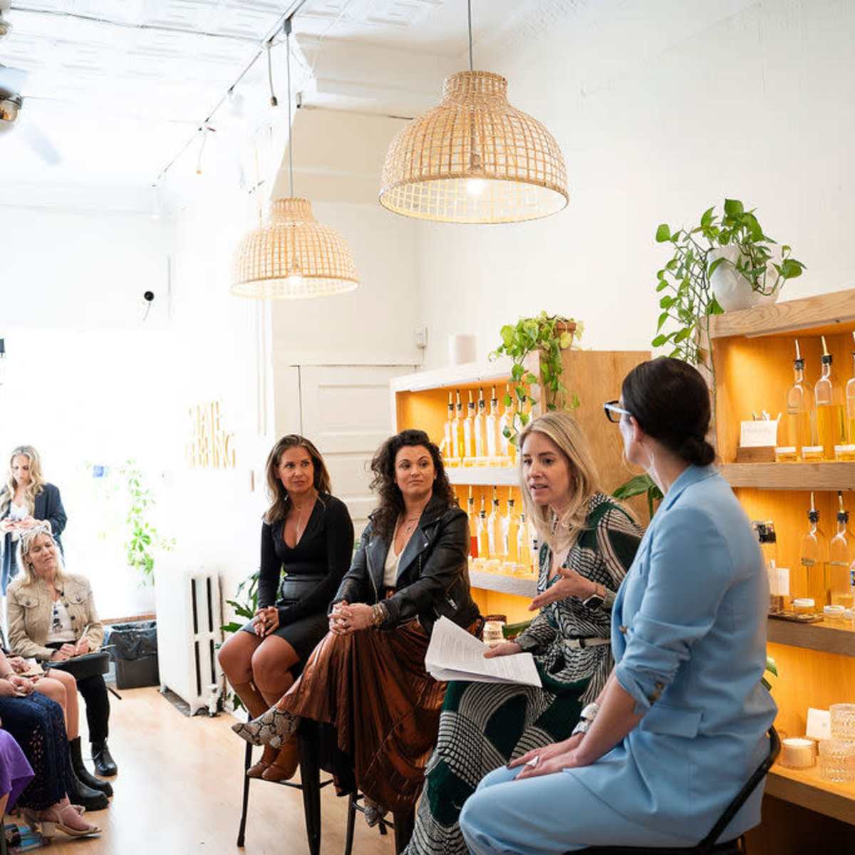 A group of women engage in a discussion in a brightly lit shop with plants and bottles displayed on the shelves, while an audience listens attentively.