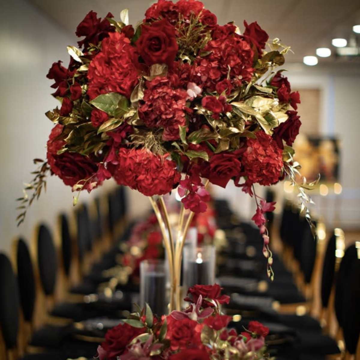A dining table is elegantly set with tall floral centerpieces made of red roses and hydrangeas, surrounded by smaller arrangements and black table settings.