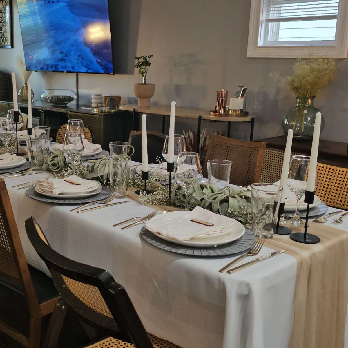 A long dining table is set for a formal meal, adorned with white tablecloths, plates, glassware, and candles, in a well-lit room with decorative elements.