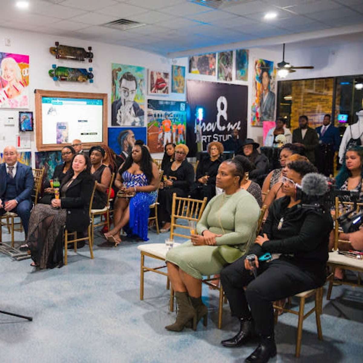 A woman speaks at a microphone in an art gallery filled with attendees seated and standing among colorful artworks on the walls.