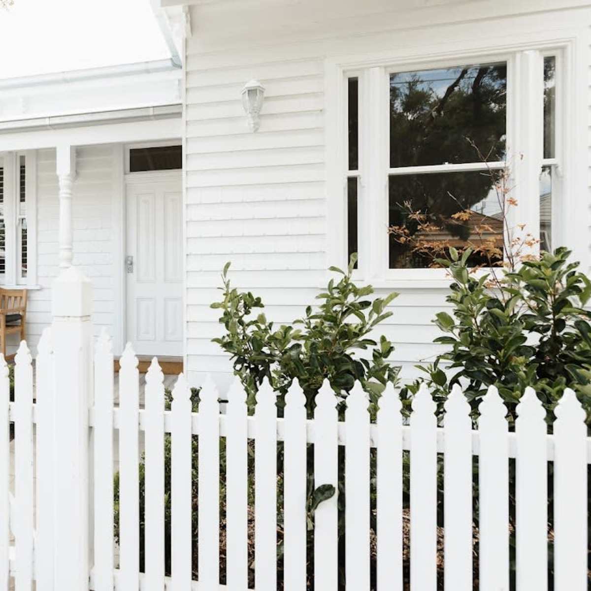A family sits on a bench on the porch of a white house surrounded by a white picket fence and green shrubs.