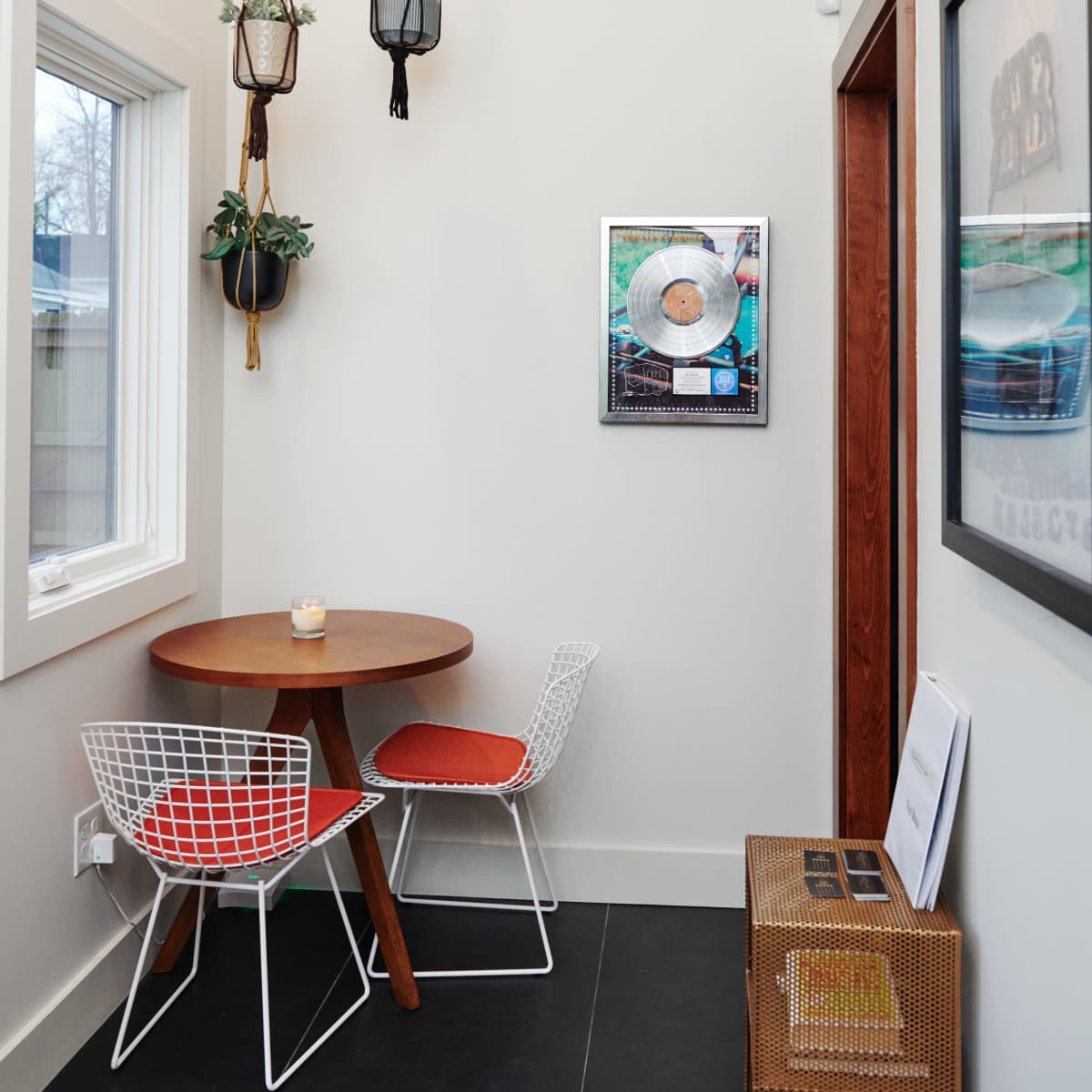 A small dining nook features a round wooden table with two wire chairs and hanging planters above, adjacent to a framed record on the wall.