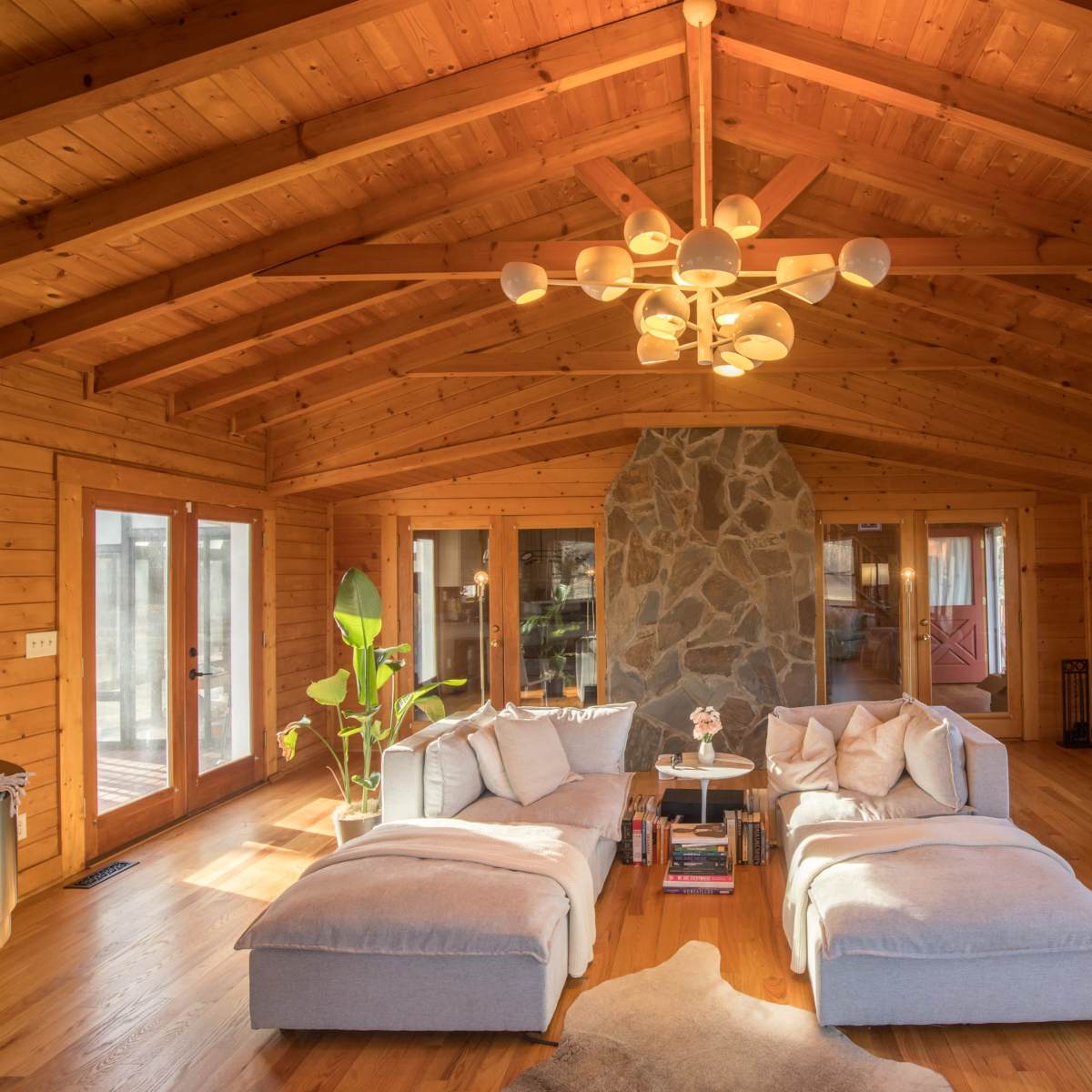 The living room features a high wooden ceiling, a stone fireplace, large windows, and two sectional sofas arranged around a central coffee table.
