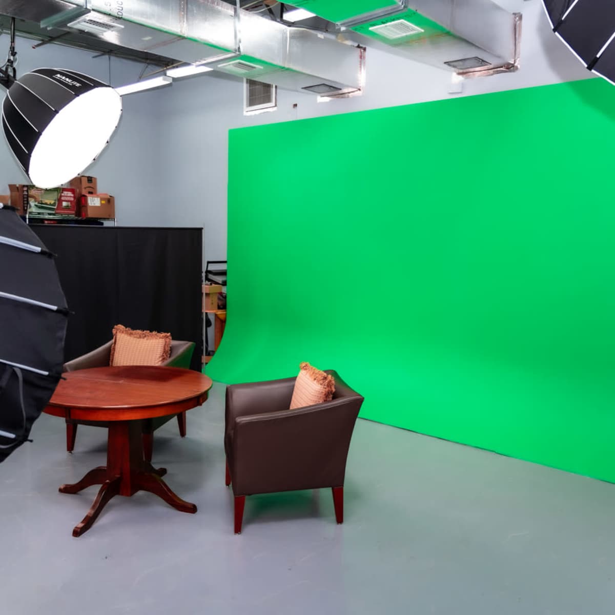A studio setup with a green backdrop, two brown chairs, a round wooden table, and several lighting equipment overhead.