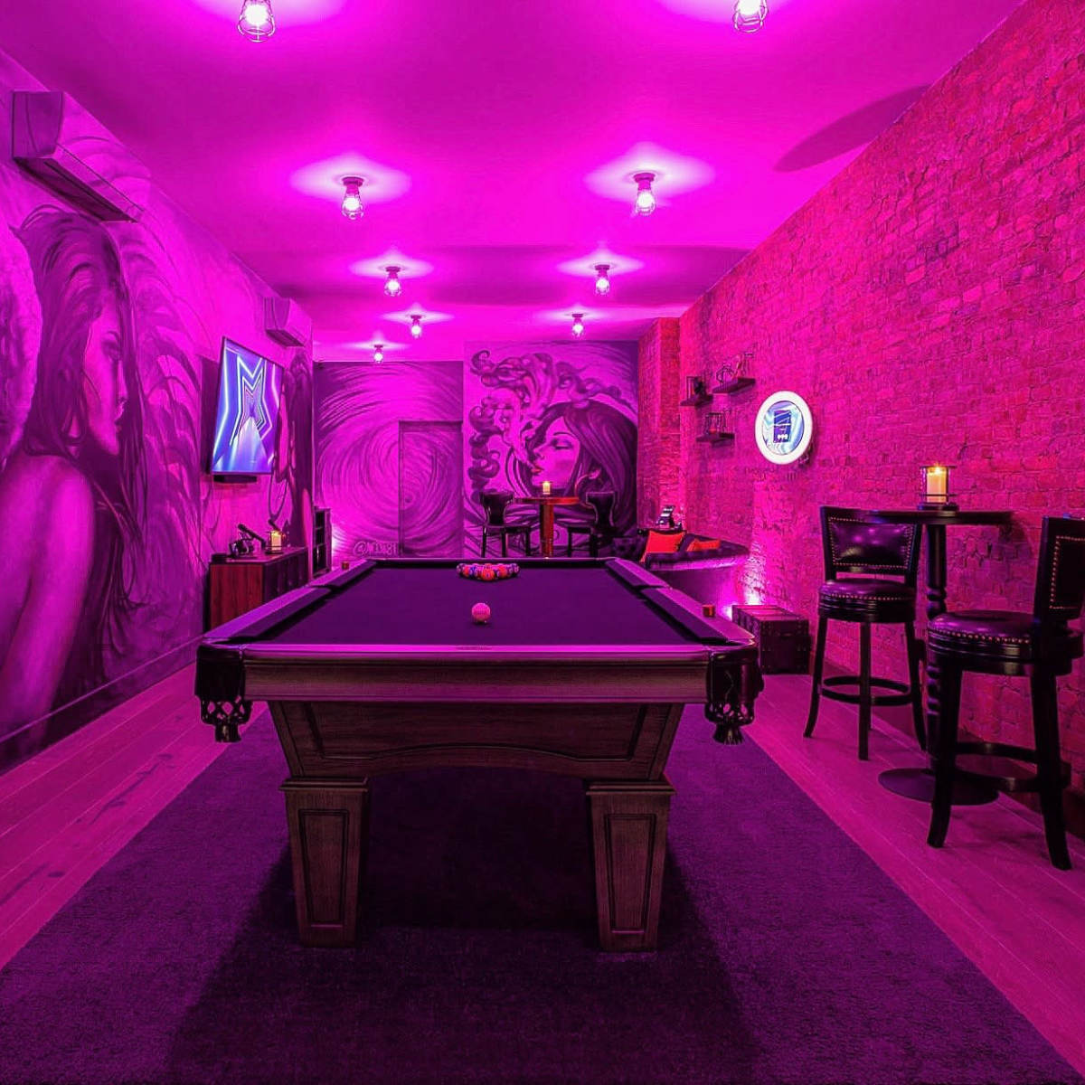 A game room features a billiards table surrounded by brick walls painted with vibrant murals and illuminated by pink lights.