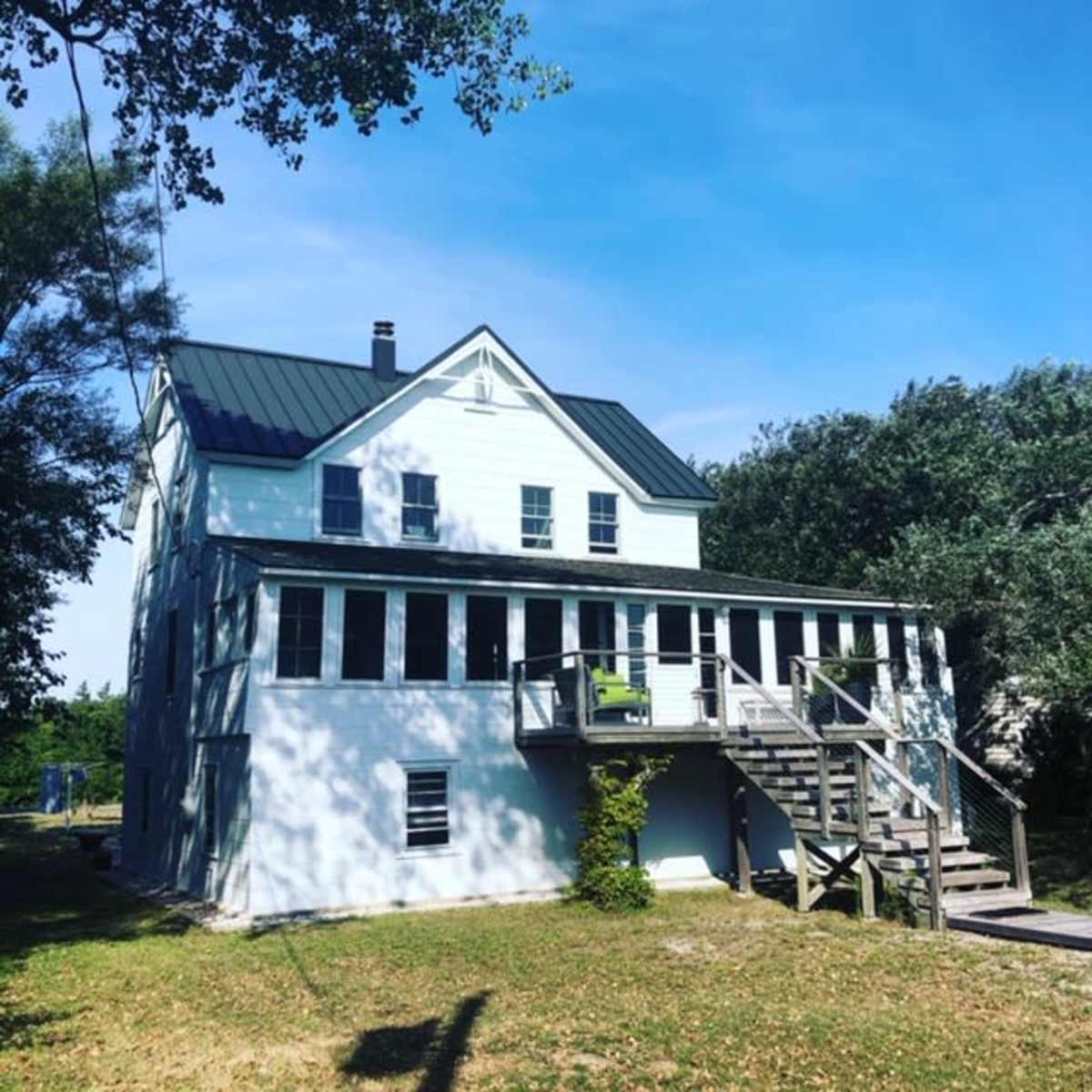 A two-story house with a metal roof features a large porch and stairs leading to the yard.