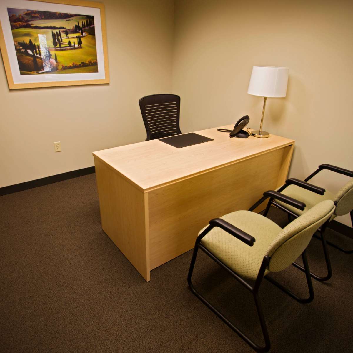 A simple office space features a desk with a phone, a lamp, and two chairs, alongside a framed landscape painting on the wall.