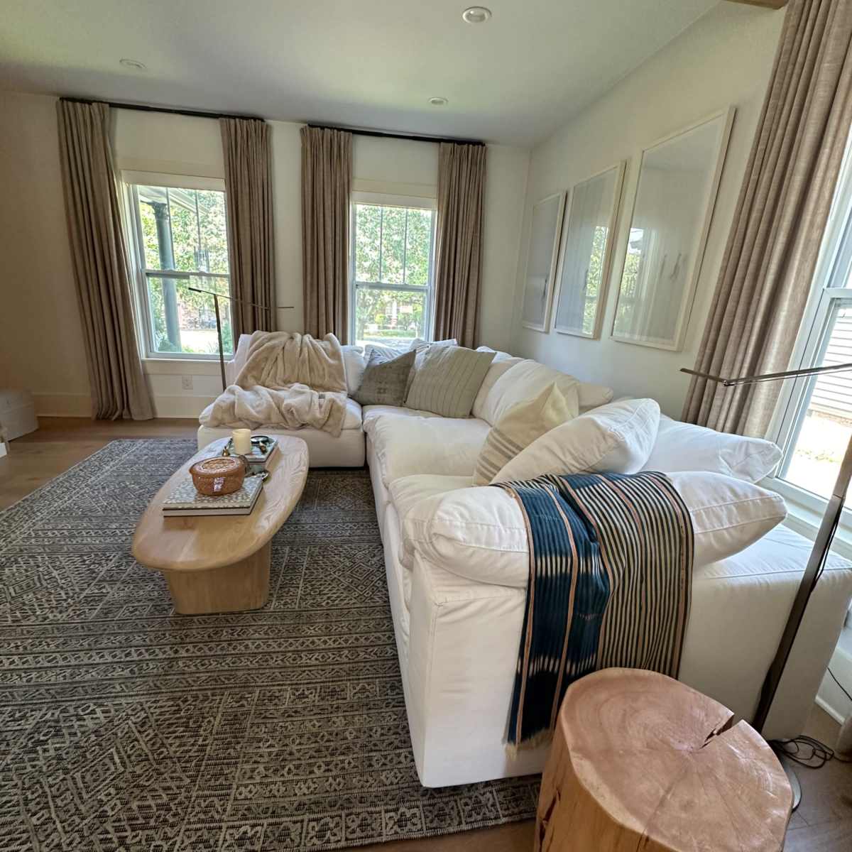 A bright living room features a large white sectional sofa, a wooden coffee table, and two tall windows adorned with curtain panels.
