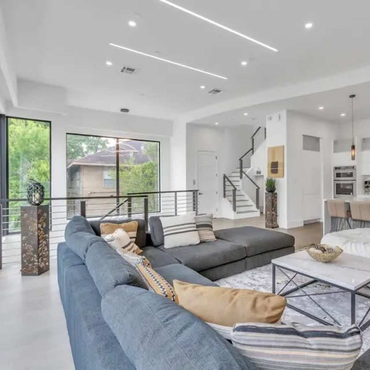 A modern living room with large windows, a sectional sofa, and a kitchen area featuring bar stools and sleek cabinetry.