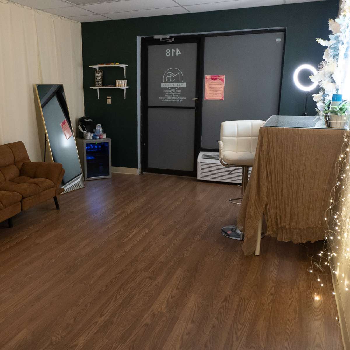 A small, softly lit room featuring a brown sofa, a mirror, a mini fridge, and a desk with decorative elements, along with a large door.