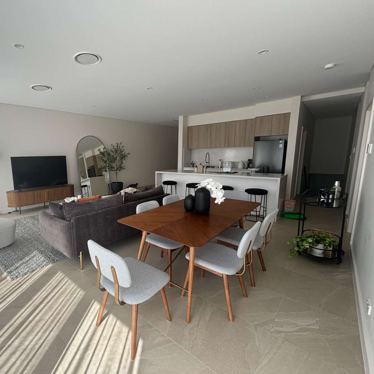 A modern open-concept living and dining area with a large dining table, a sofa, a television, and a kitchen in the background.
