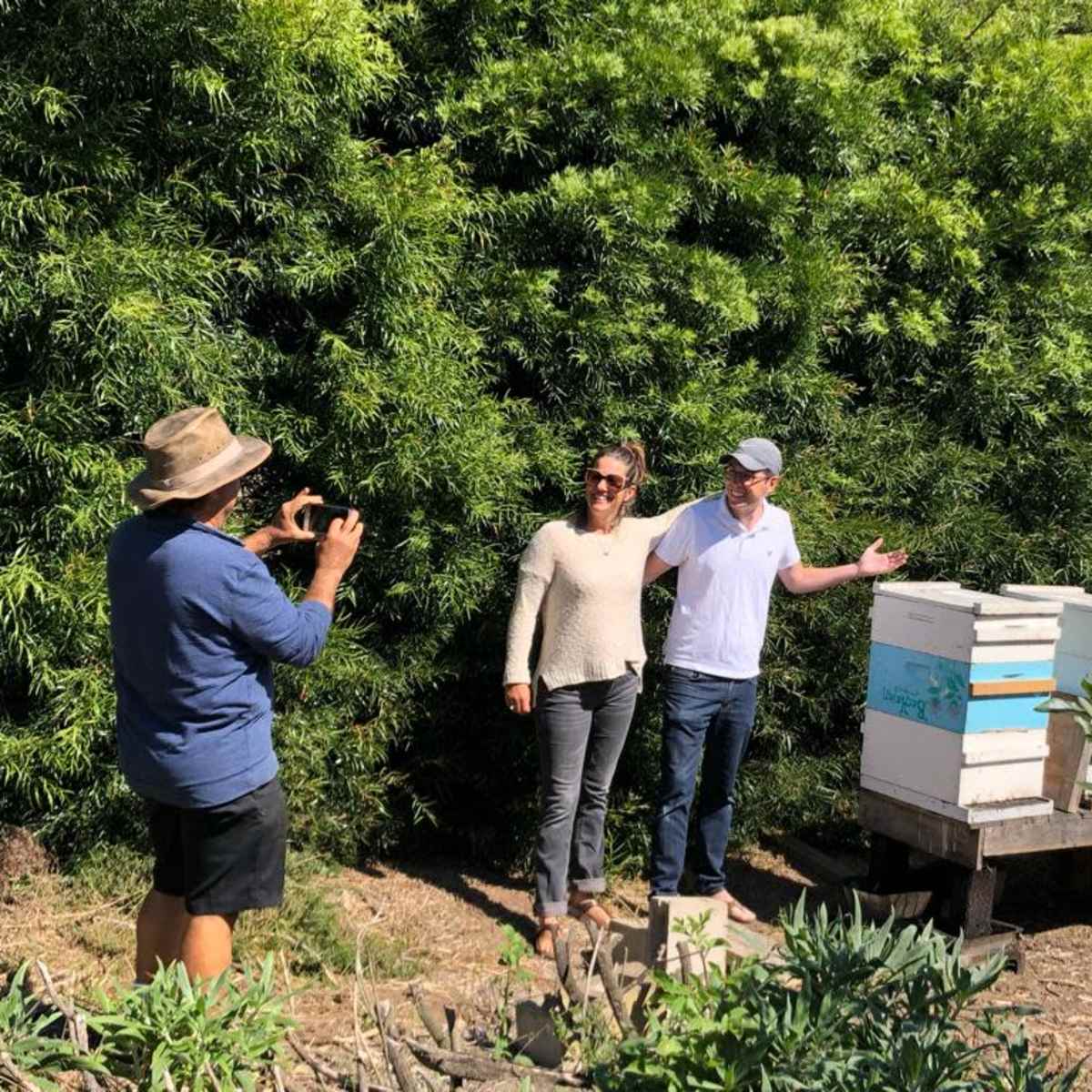 Two people stand beside a row of beehives while a third person photographs them in a garden setting.
