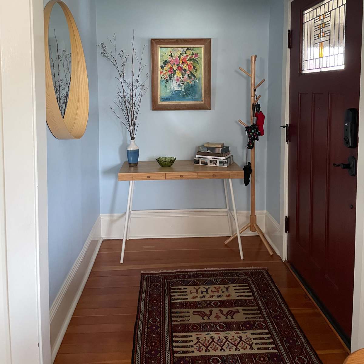A bright hallway with a blue wall, featuring a decorative mirror, a framed painting, a small table, a plant, and a coat rack, alongside a dark-colored front door.