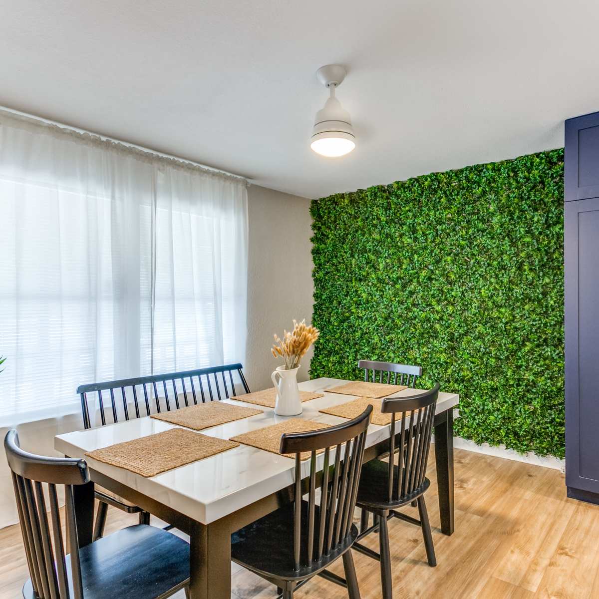 A dining area features a table with black chairs, a vase of dried flowers, and a green wall of artificial plants, alongside a dark blue cabinet and a large window with sheer curtains.