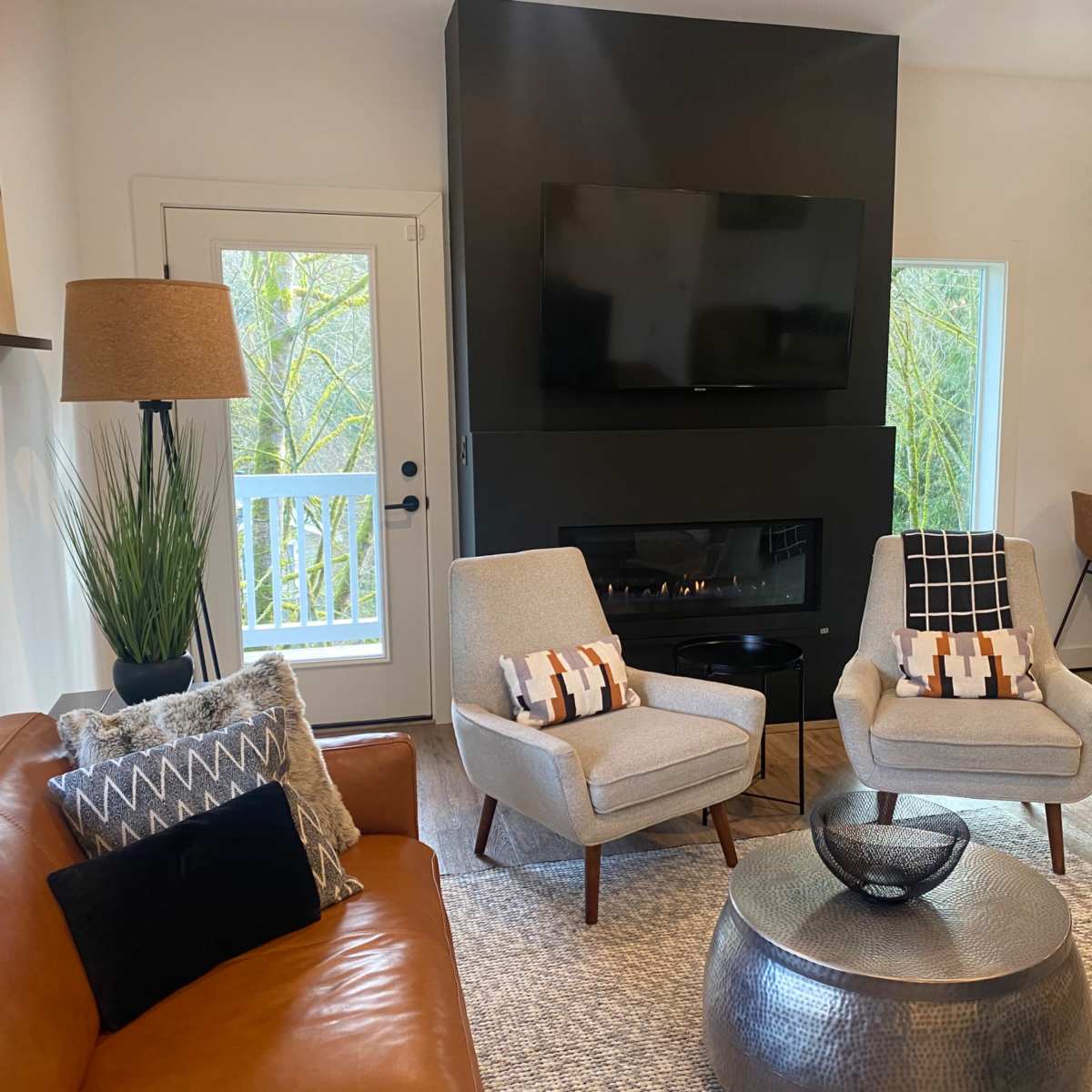 A modern living room with a brown leather sofa, two light gray chairs, a round metallic coffee table, and a wall-mounted television above a fireplace.