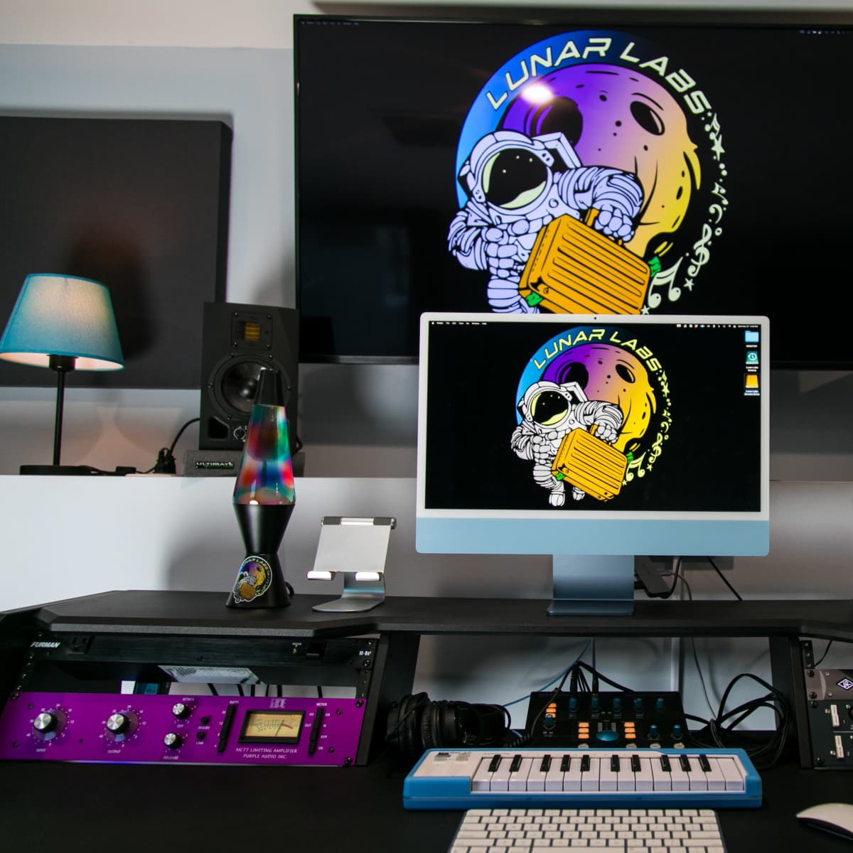 A music studio setup features a computer monitor displaying the "Lunar Labs" logo, surrounded by audio equipment, speakers, and colorful lighting.