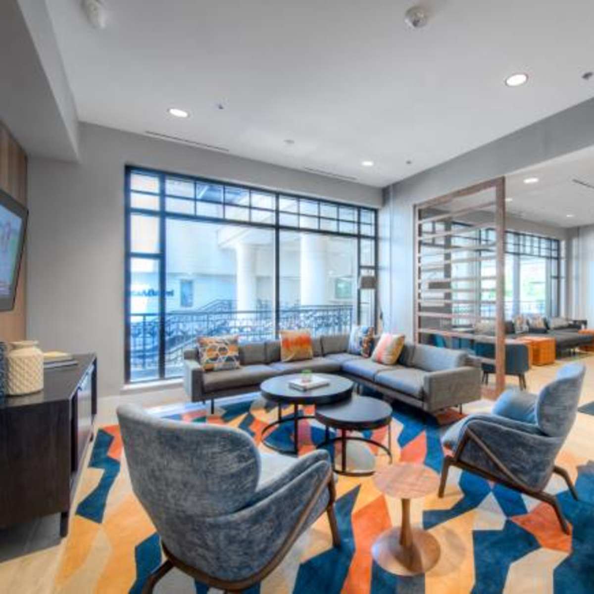 A modern lounge area features comfortable seating, a large rug with a geometric pattern, and a dining space with a round table surrounded by chairs.