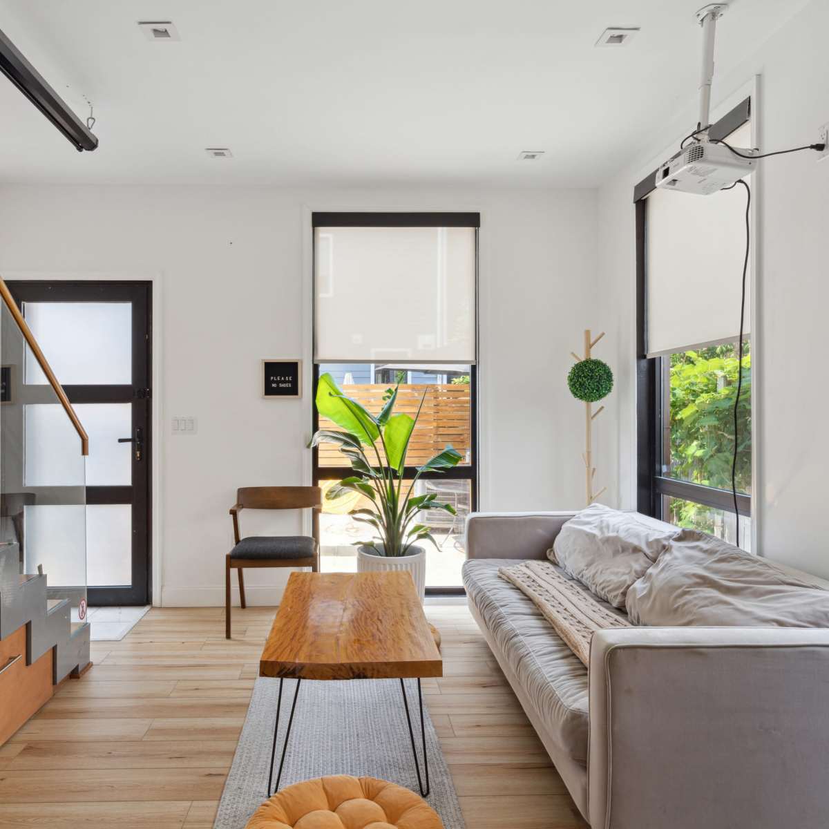 A modern living room featuring a staircase, a coffee table, a sofa, and a potted plant, with large windows allowing natural light to enter.