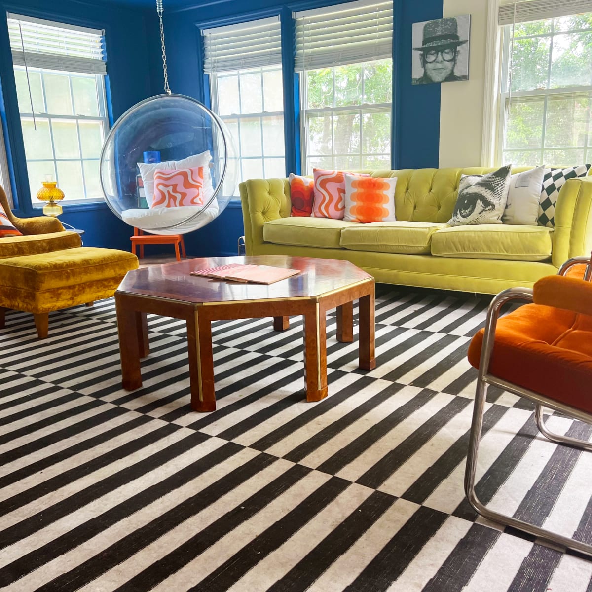 The room features a bright green sofa, an orange accent chair, a black and white striped rug, and a round glass bubble chair, creating a vibrant living space.
