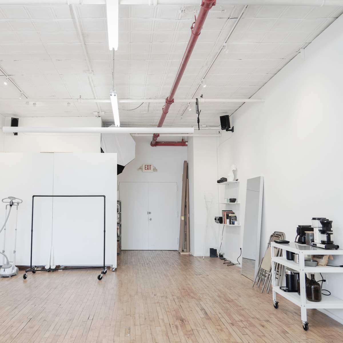 A bright, empty studio space with wooden flooring, white walls, and minimal furnishings, including a mobile partition and a small cart with kitchen equipment.