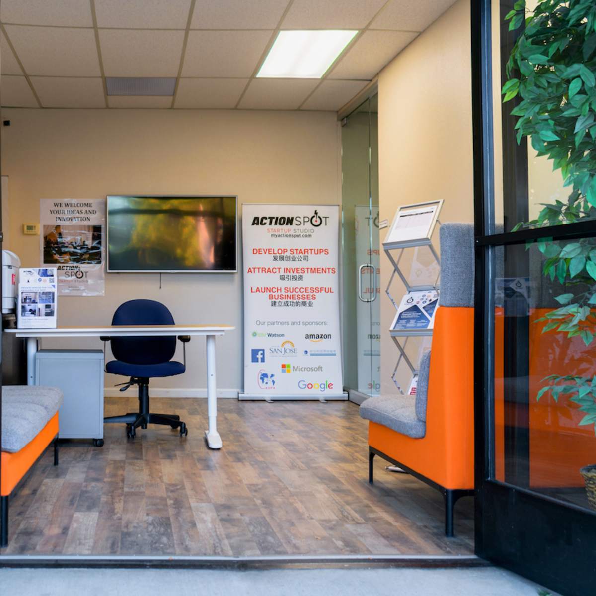 The entrance of a co-working space features a reception area with seating, a desk, and a TV displaying promotional materials.