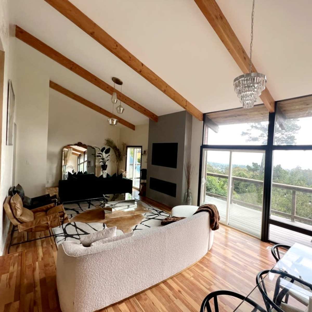 A modern living room with wooden beams, large windows overlooking a natural landscape, and minimalist furniture including a white sofa and a glass dining table.