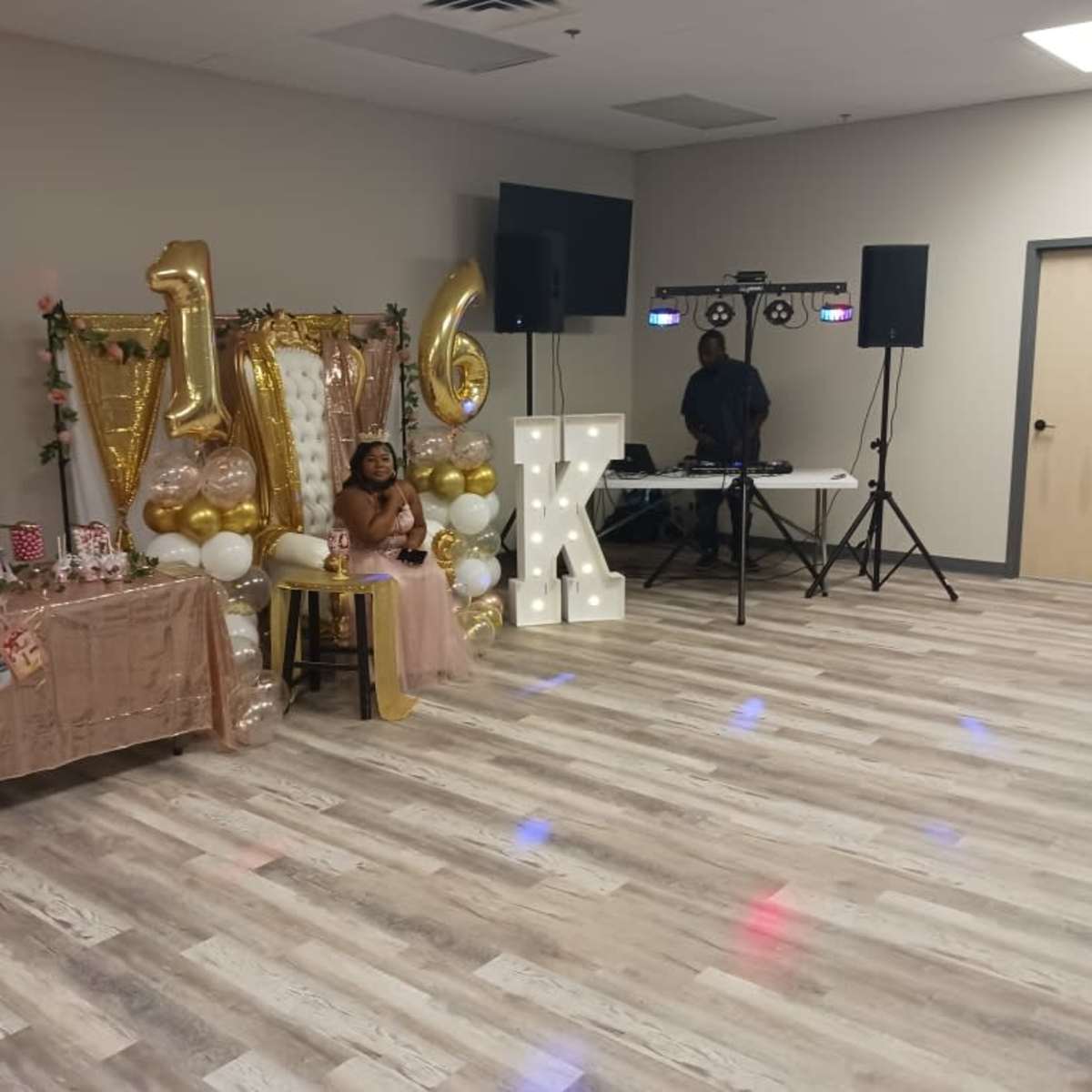 A child sits on a chair in front of a decorated table with a "16" sign and party supplies in a reception hall.