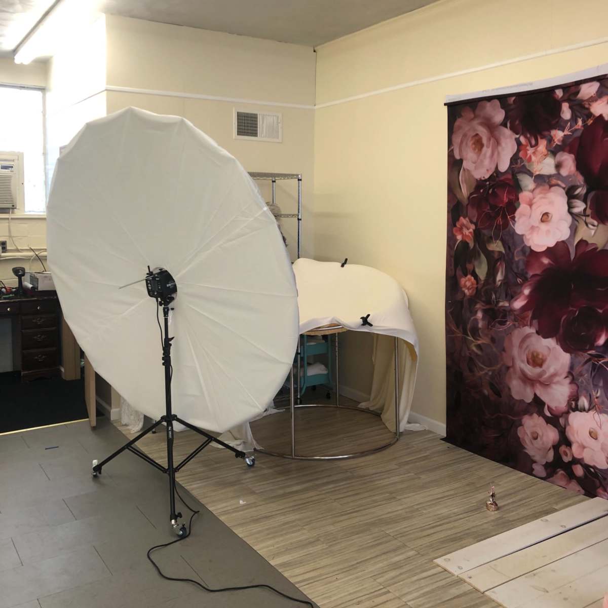 A photography studio setup featuring a large reflective umbrella light, a curved display rack covered with fabric, and a floral backdrop.