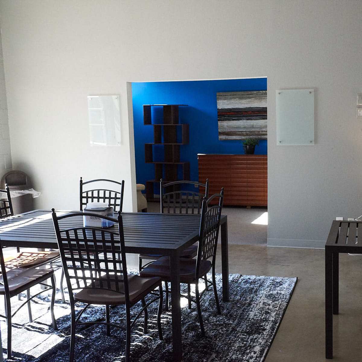 A bright, minimalist room features a dining table, chairs, and a small lamp, with blue walls and wooden furniture in the background.