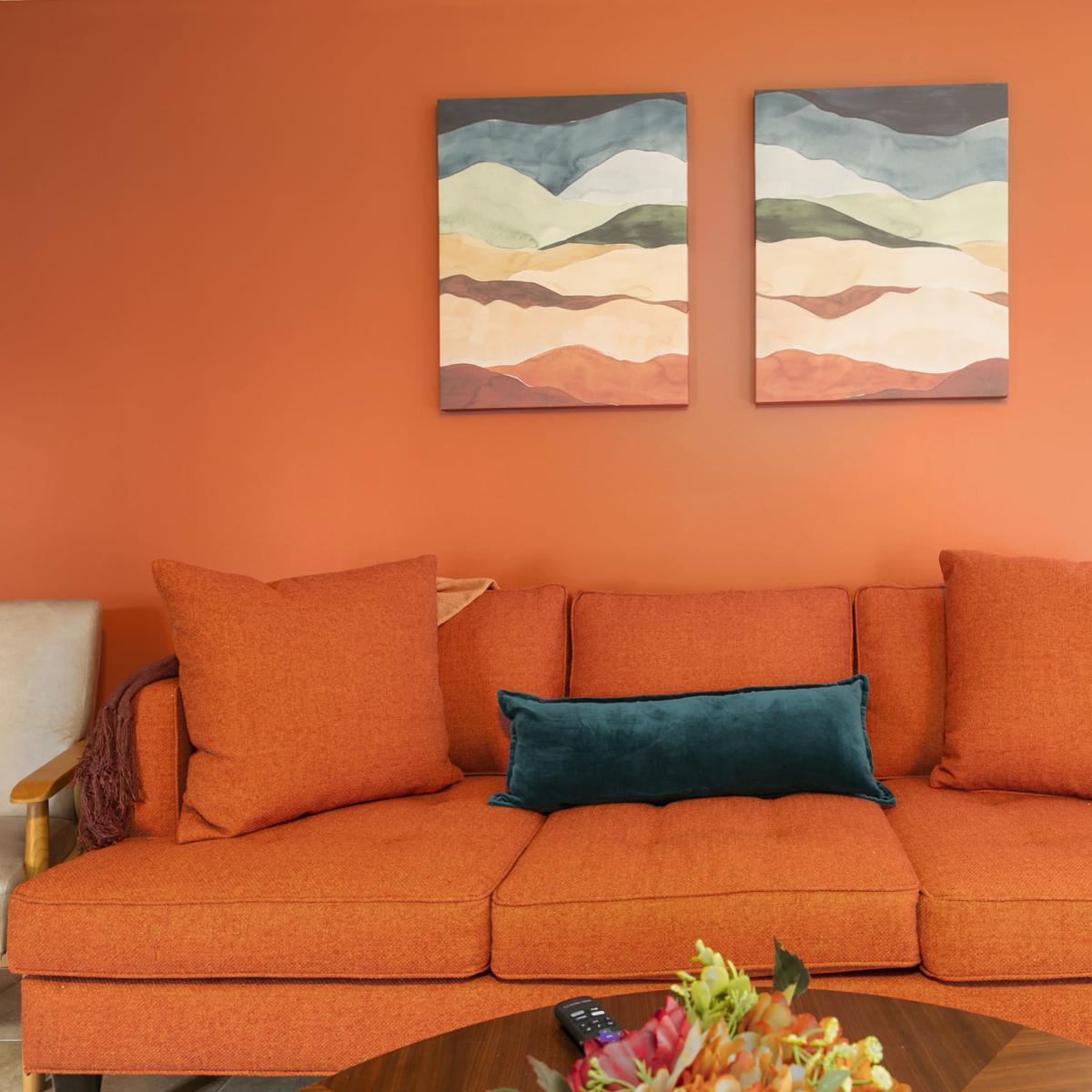 A rust-colored sofa with large cushions is positioned against a bright orange wall, adorned with two abstract paintings, while a round coffee table with a floral arrangement sits in front.