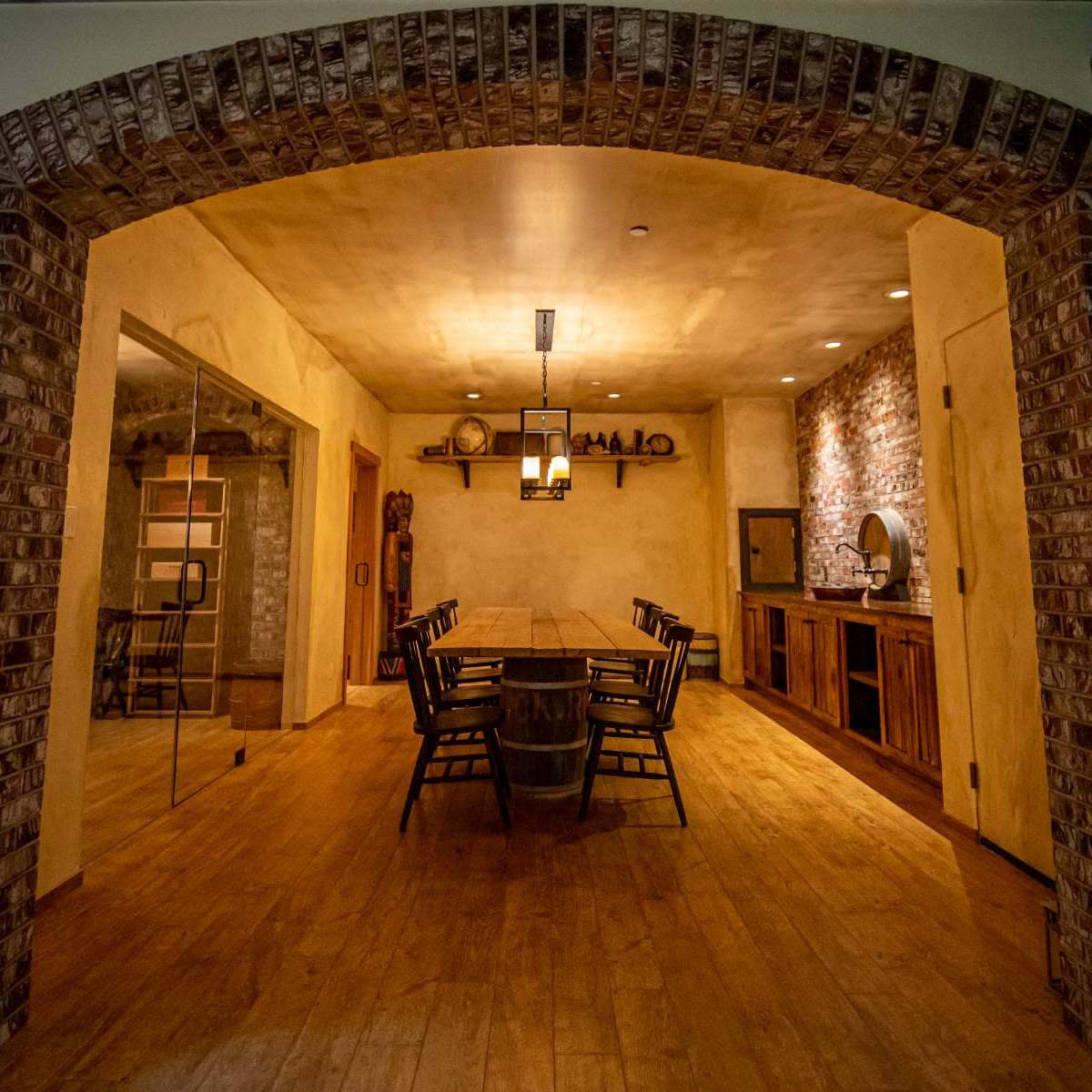 A warm, rustic dining room with wooden flooring, a long table surrounded by chairs, and a brick archway leading to another area.
