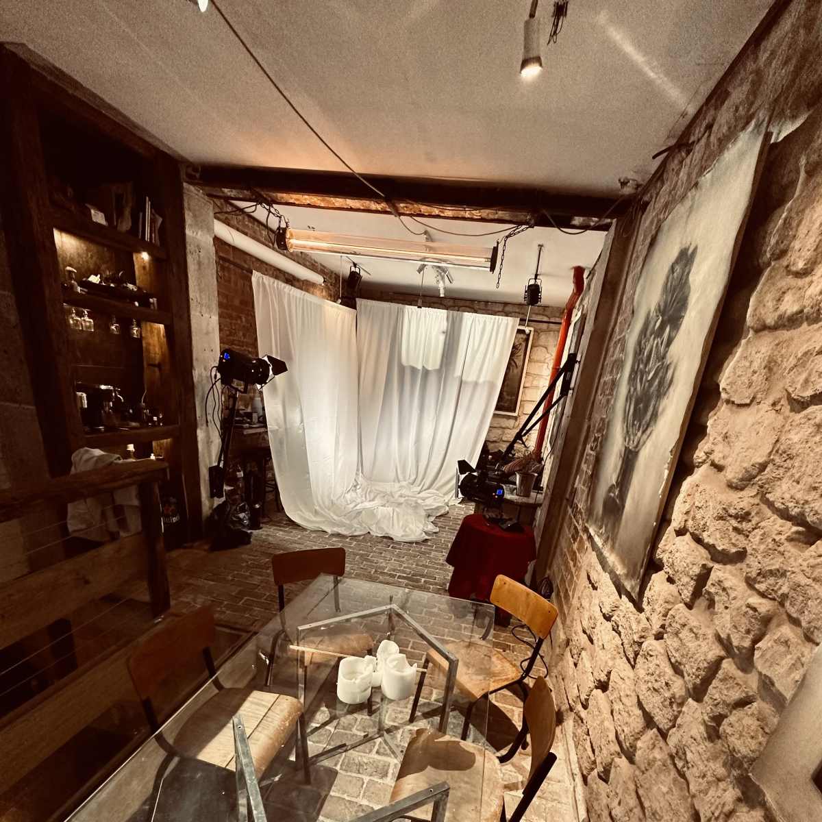 A glass-topped table with wooden chairs is set in a stone-walled room, featuring equipment and backdrops for filming in the background.