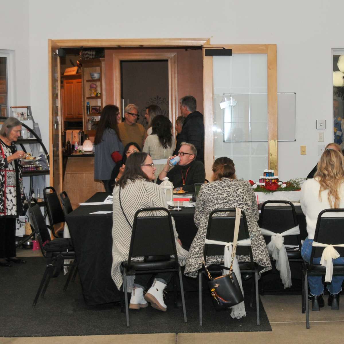 A group of people are gathered around a table in a lobby area, while others are entering and exiting through a nearby door.