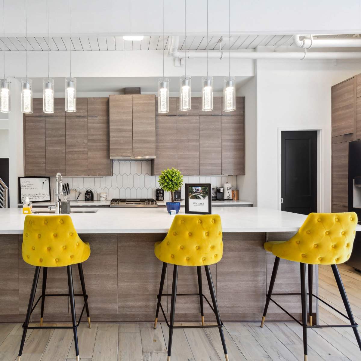 A modern kitchen features a large island with four yellow upholstered bar stools, pendant lights hanging above, and wooden cabinetry along the walls.