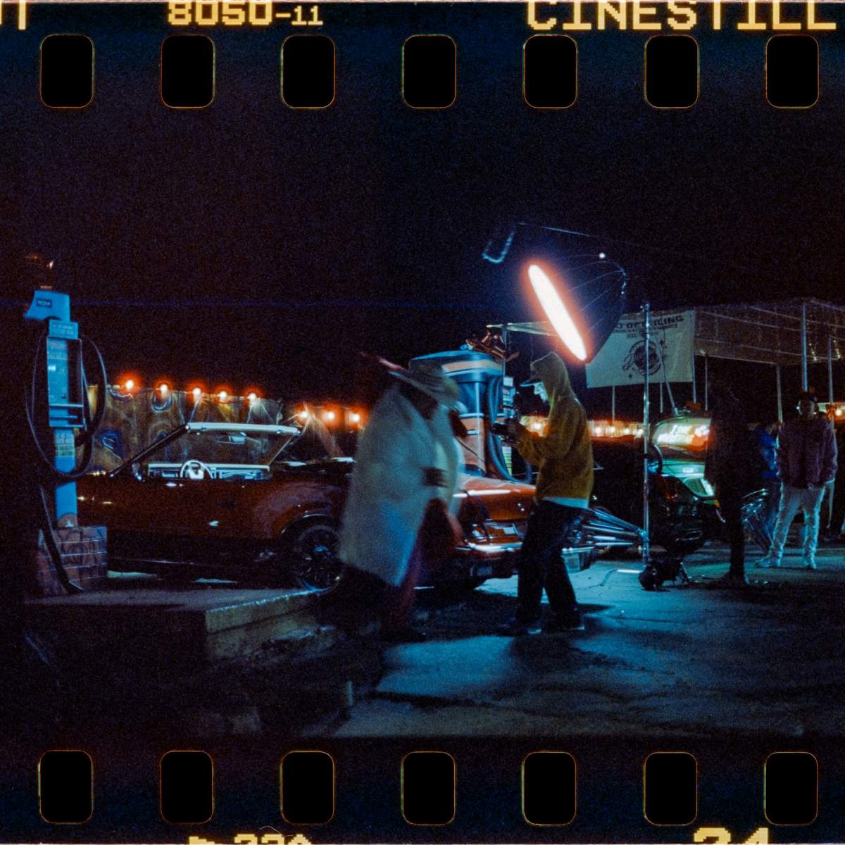 The scene depicts a nighttime shooting location with cars, crew members, and lighting equipment set up for a film or photo shoot.