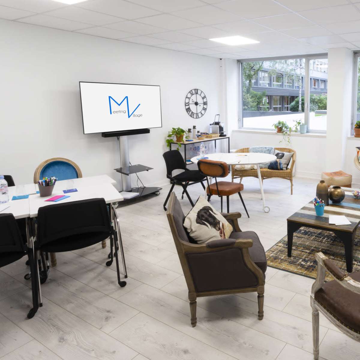 A bright office space featuring a meeting area with a large screen, several seating arrangements, and windows that provide views of greenery outside.