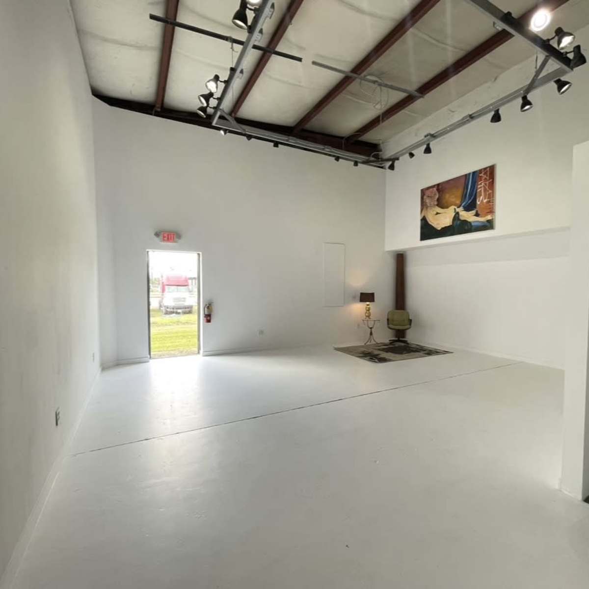 An empty room with white walls, a door leading outside, a lamp, a chair, and an artwork on the wall.