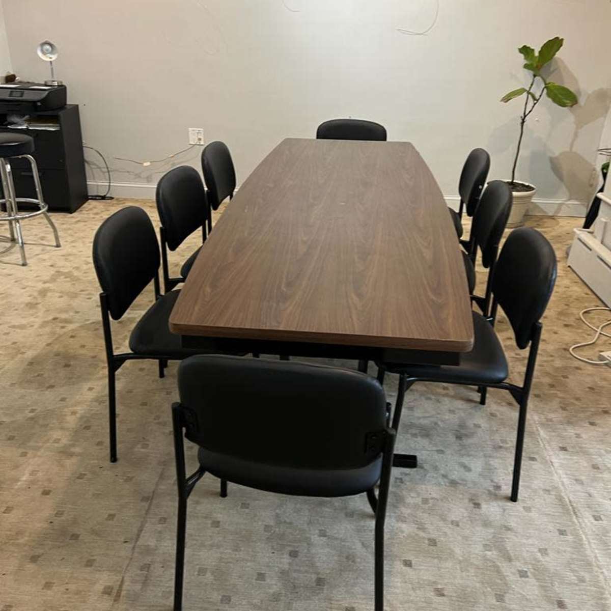 A rectangular wooden table surrounded by eight black chairs is set in an empty room with a light-colored carpet and a small plant in the corner.