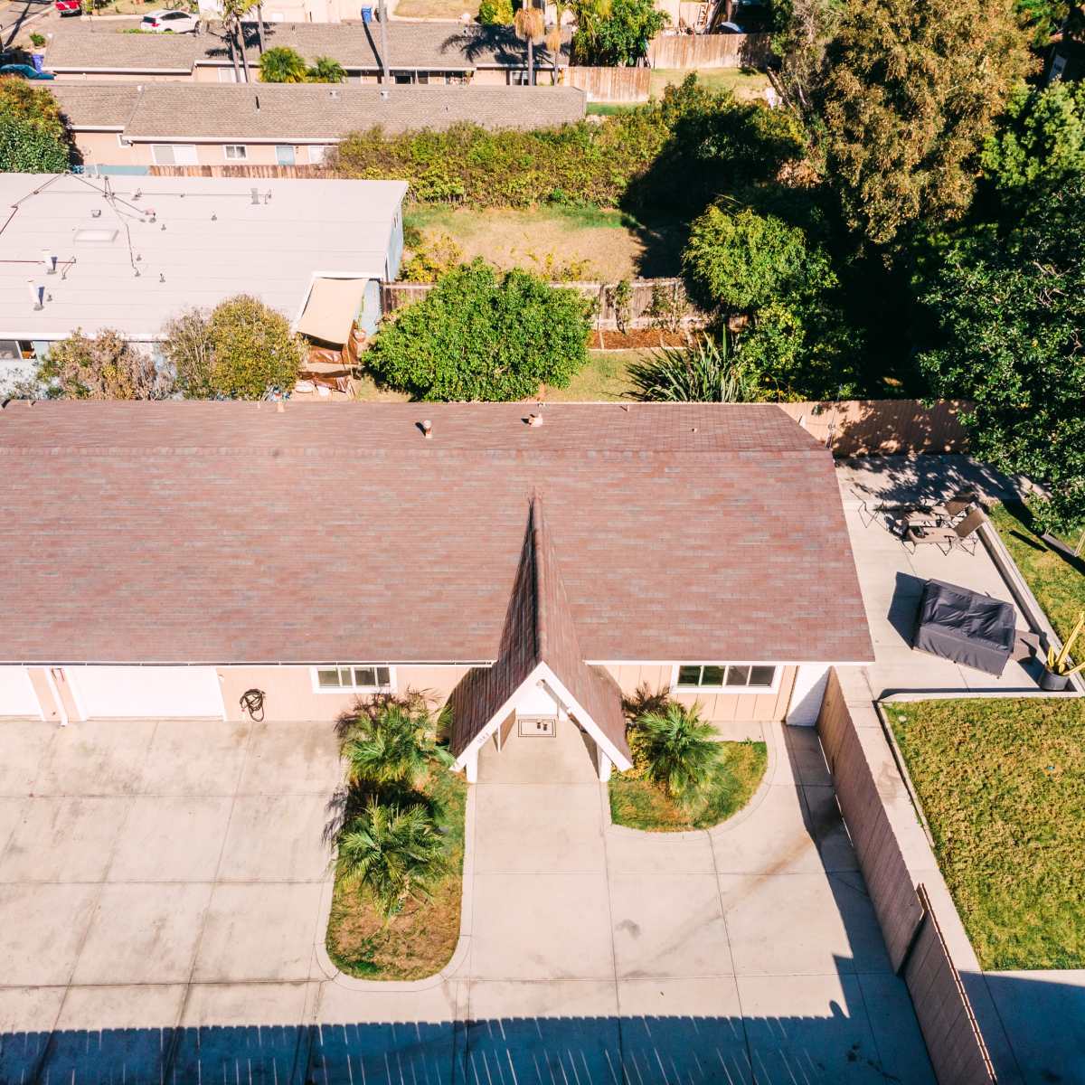 An aerial view of a residential property with a pitched roof, surrounded by greenery and adjacent structures in a suburban area.