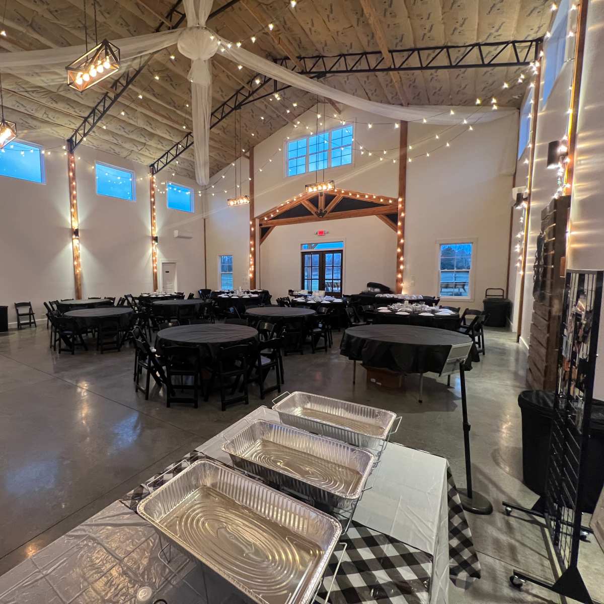 A spacious event hall with round black tables set for dining, string lights overhead, and a service table with aluminum trays in the foreground.