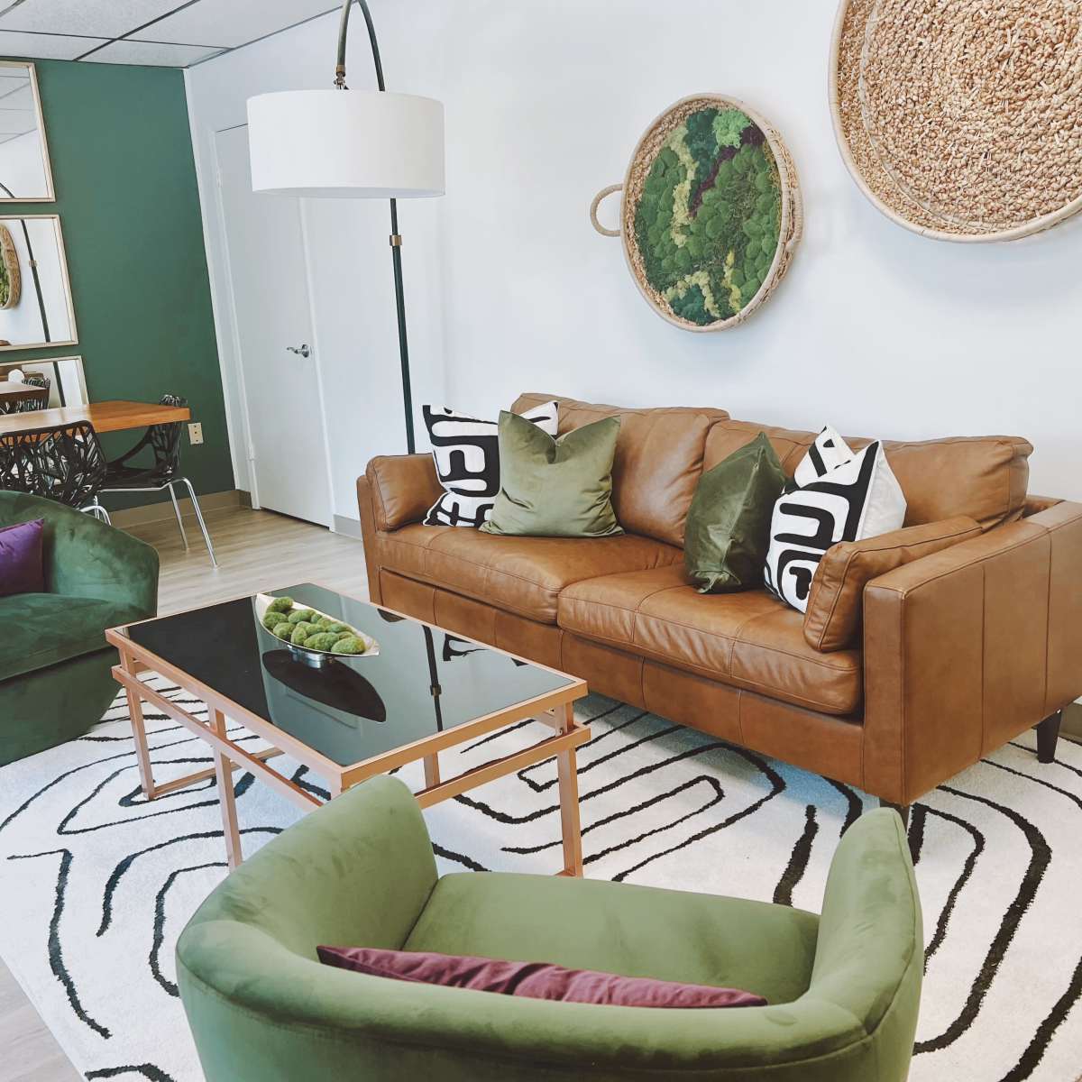 A stylish living room with a brown leather sofa, two green chairs, a glass coffee table, and wall decor featuring woven baskets.