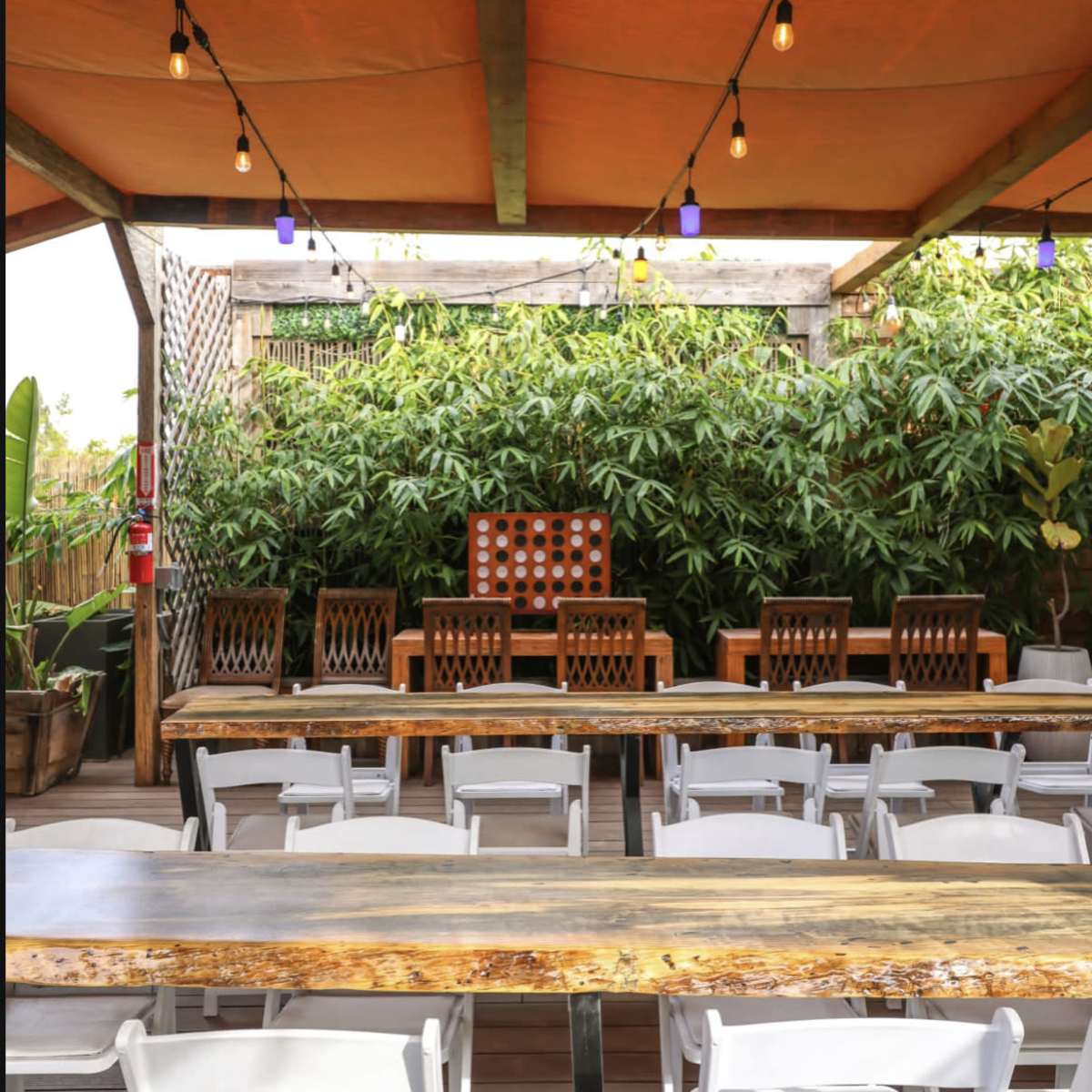 An outdoor dining area with a long wooden table, white chairs arranged in rows, and a backdrop of lush greenery.