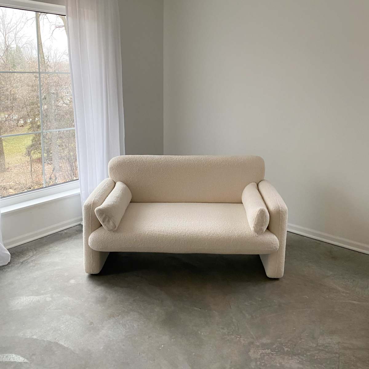 A beige, plush sofa with rounded arms sits in a minimalist room featuring a large window with sheer white curtains.
