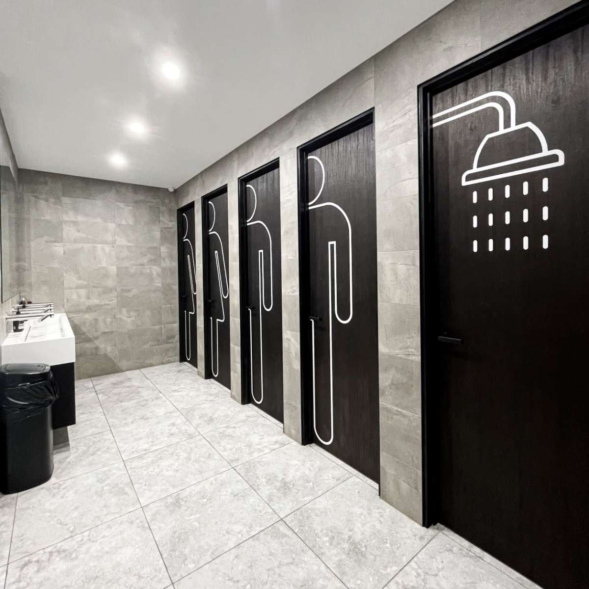 A modern restroom features four doors with minimalist symbols indicating men's and women's facilities and a shower area, alongside a sink and trash bin.