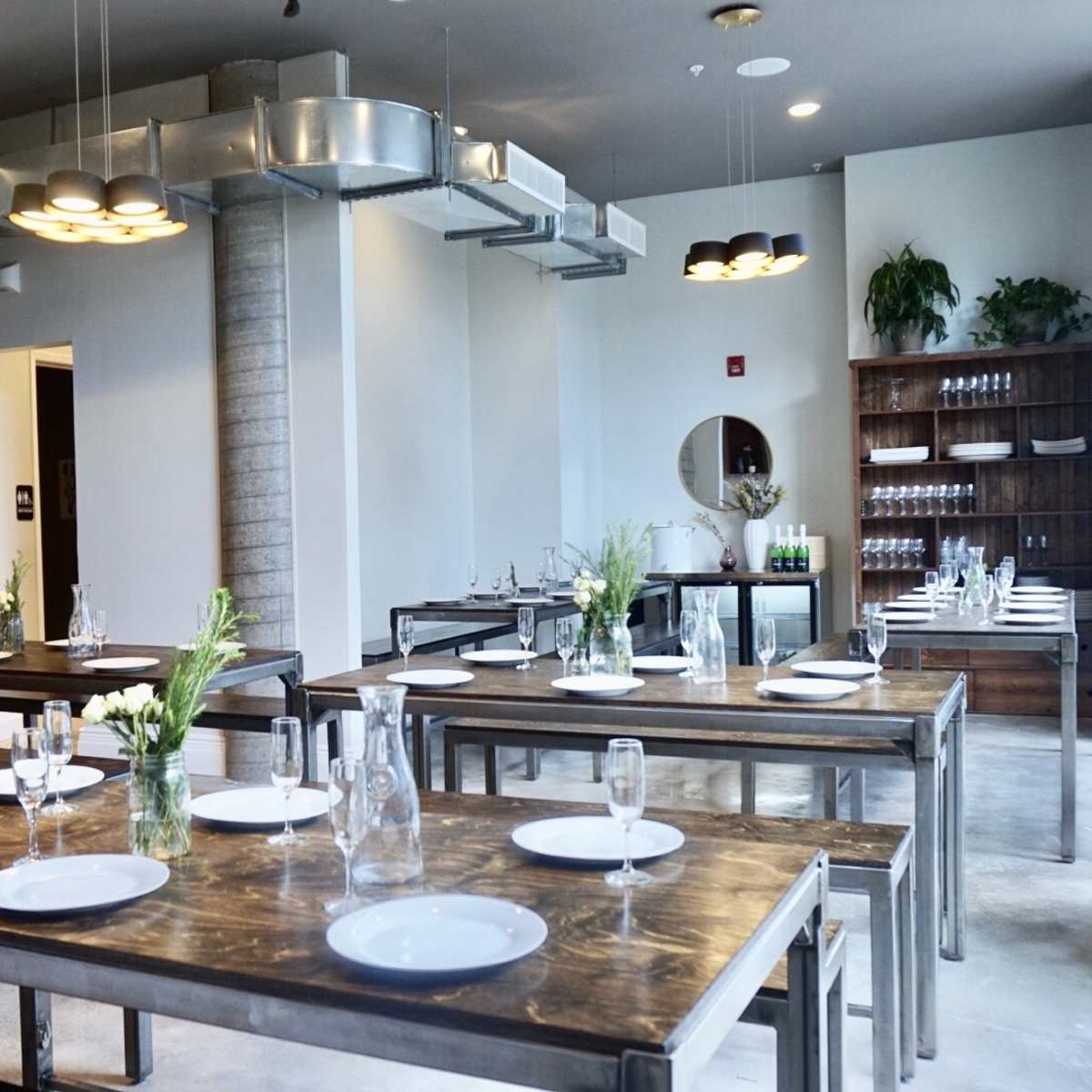 A modern dining area with several long wooden tables, each set with white plates and vases of flowers, and a wall lined with shelves holding glassware.